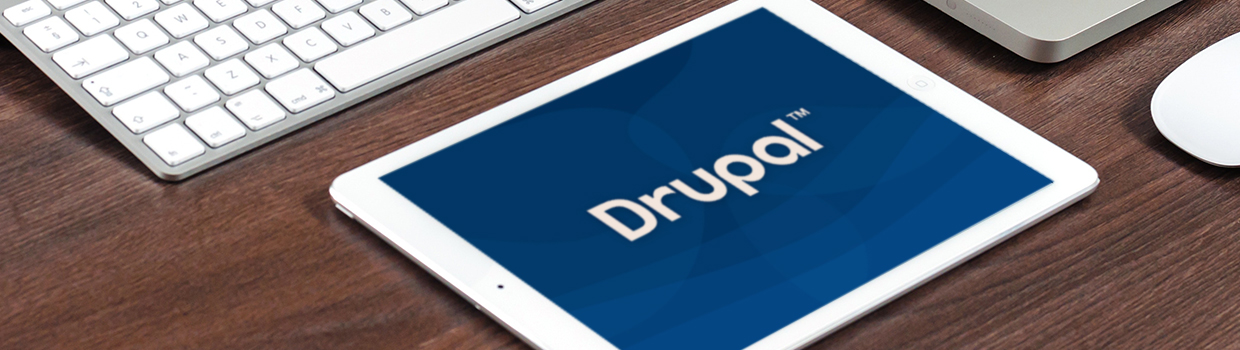 tablet with the Drupal logo displayed on it sitting on a wood grained desk next to a Mac keyboard, mouse, and laptop