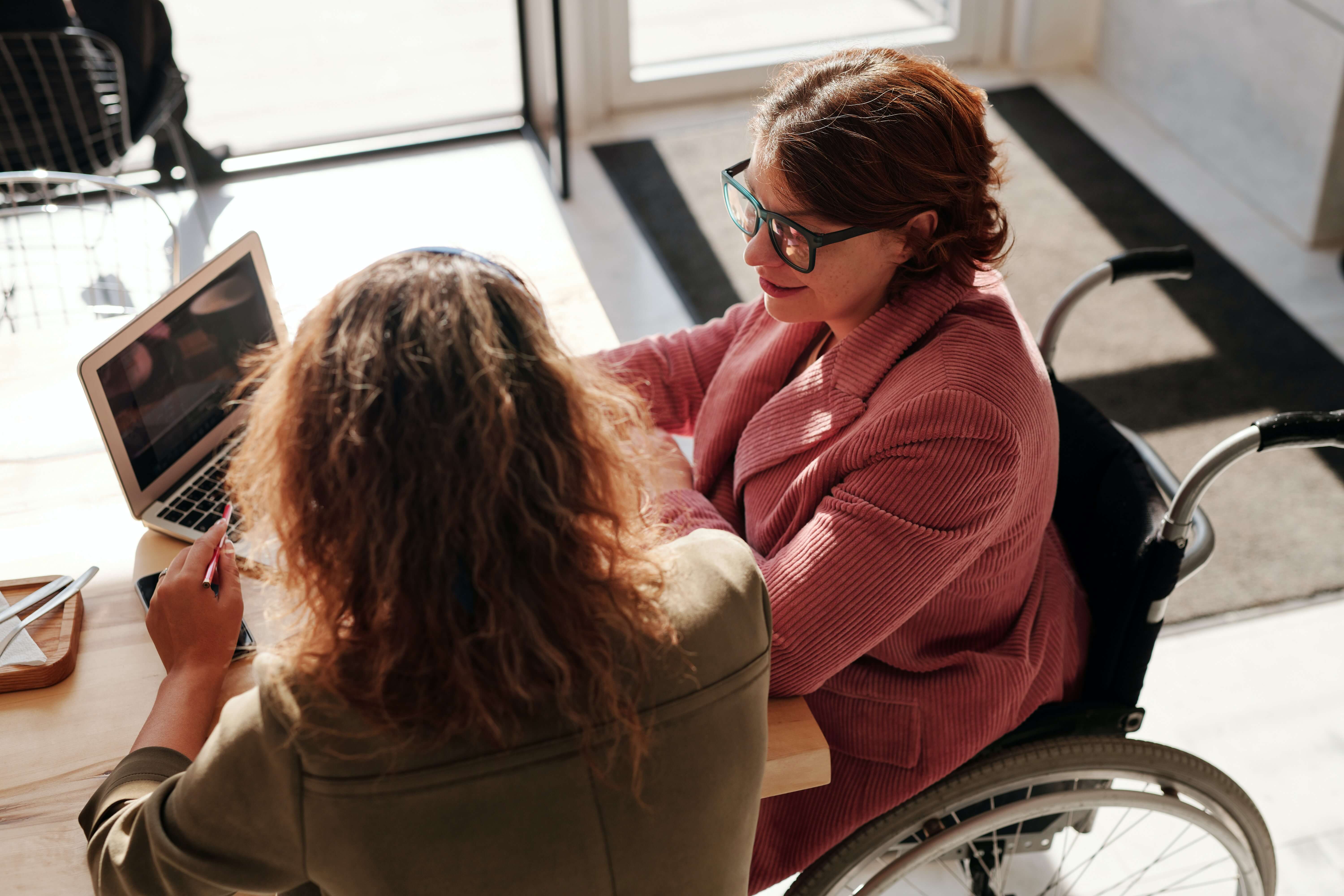 One woman in a wheelchair discusses medical results with another woman while looking at a laptop seated at a table.