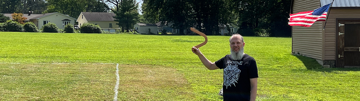 Banner image of Scott Perry outside in a field smiling at the camera and holding up a boomerang