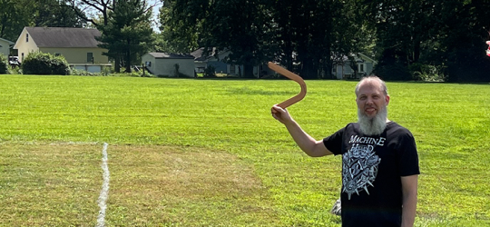 Banner image of Scott Perry outside in a field smiling at the camera and holding up a boomerang